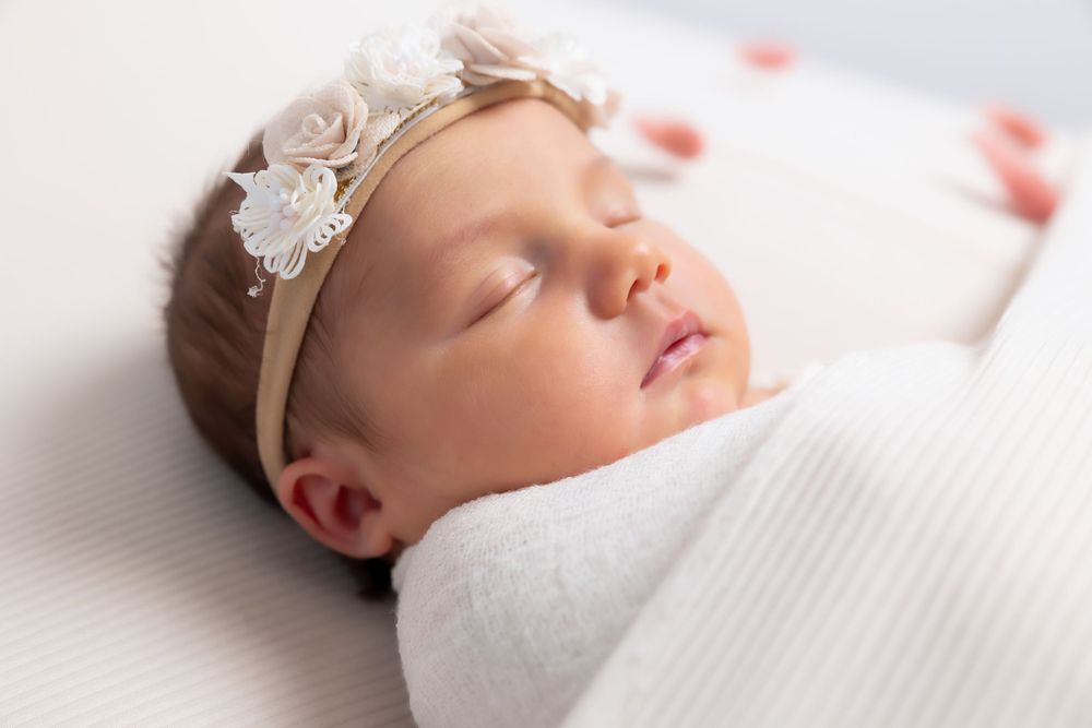 Sleeping newborn baby wearing white lace headband wrapped in soft white swaddle blanket.