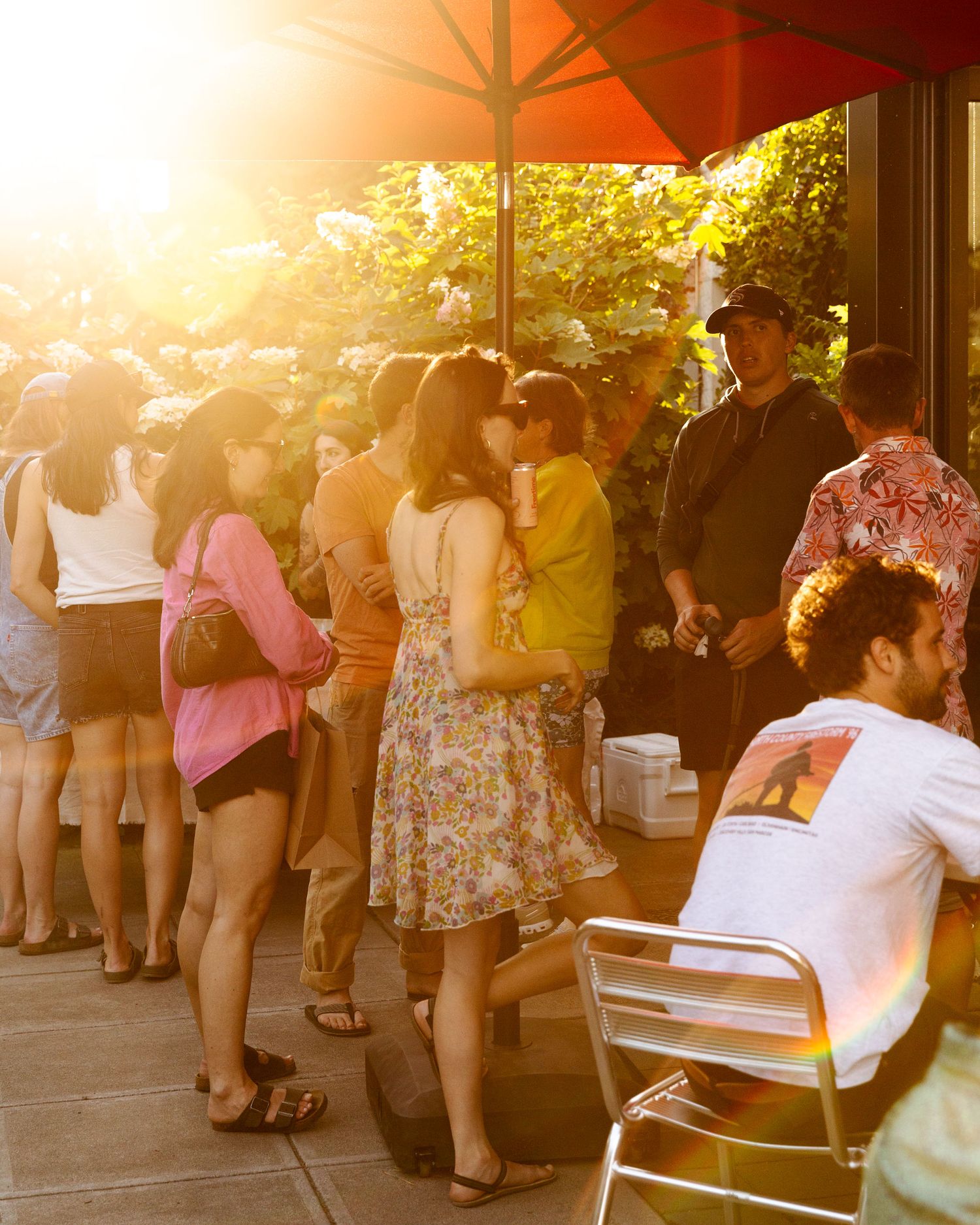 A crowd of people wait in line under an orange umbrella during golden hour at an outdoor venue.