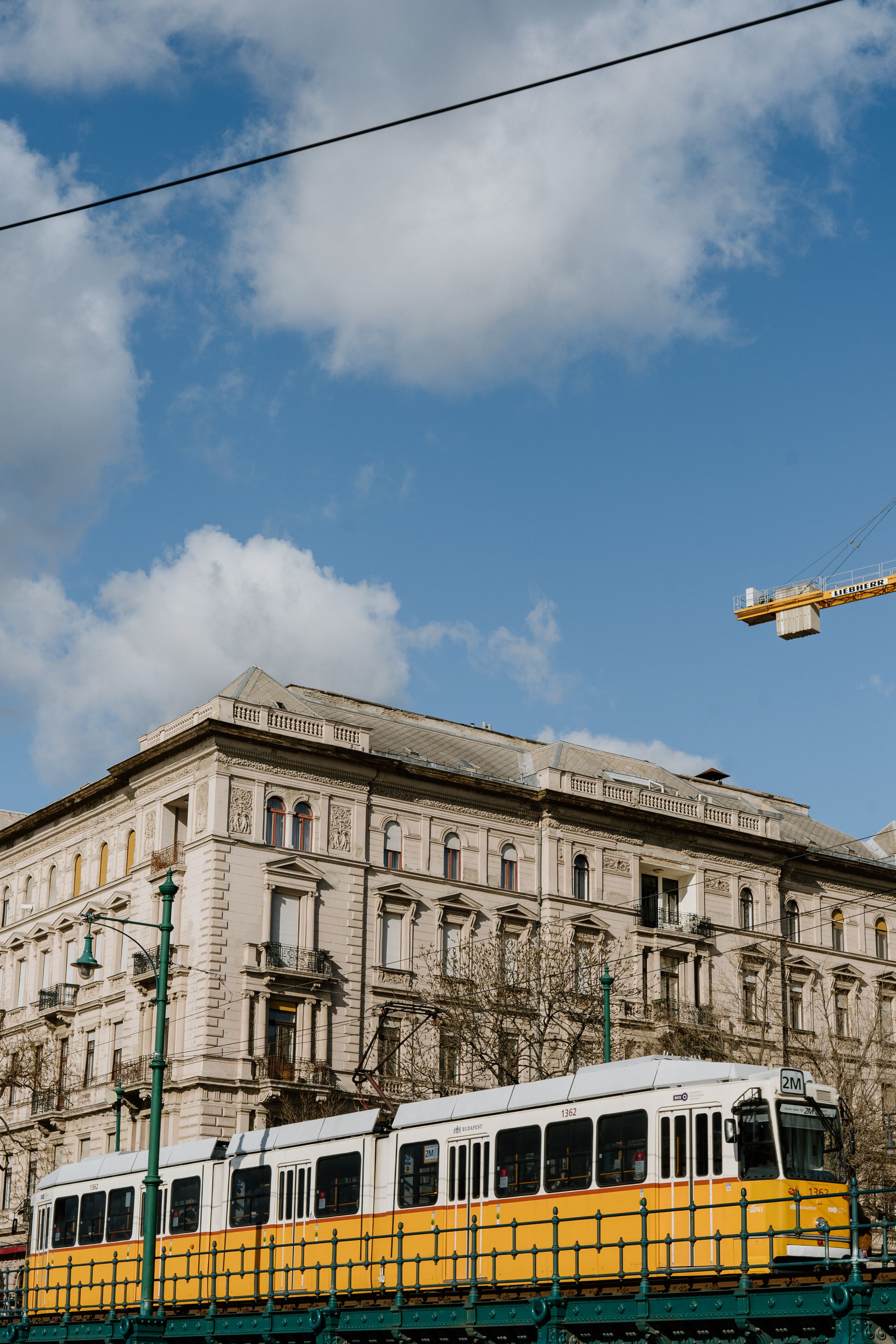 Yellow tram passes by historic stone building in Budapest with construction crane visible against blue cloudy sky.