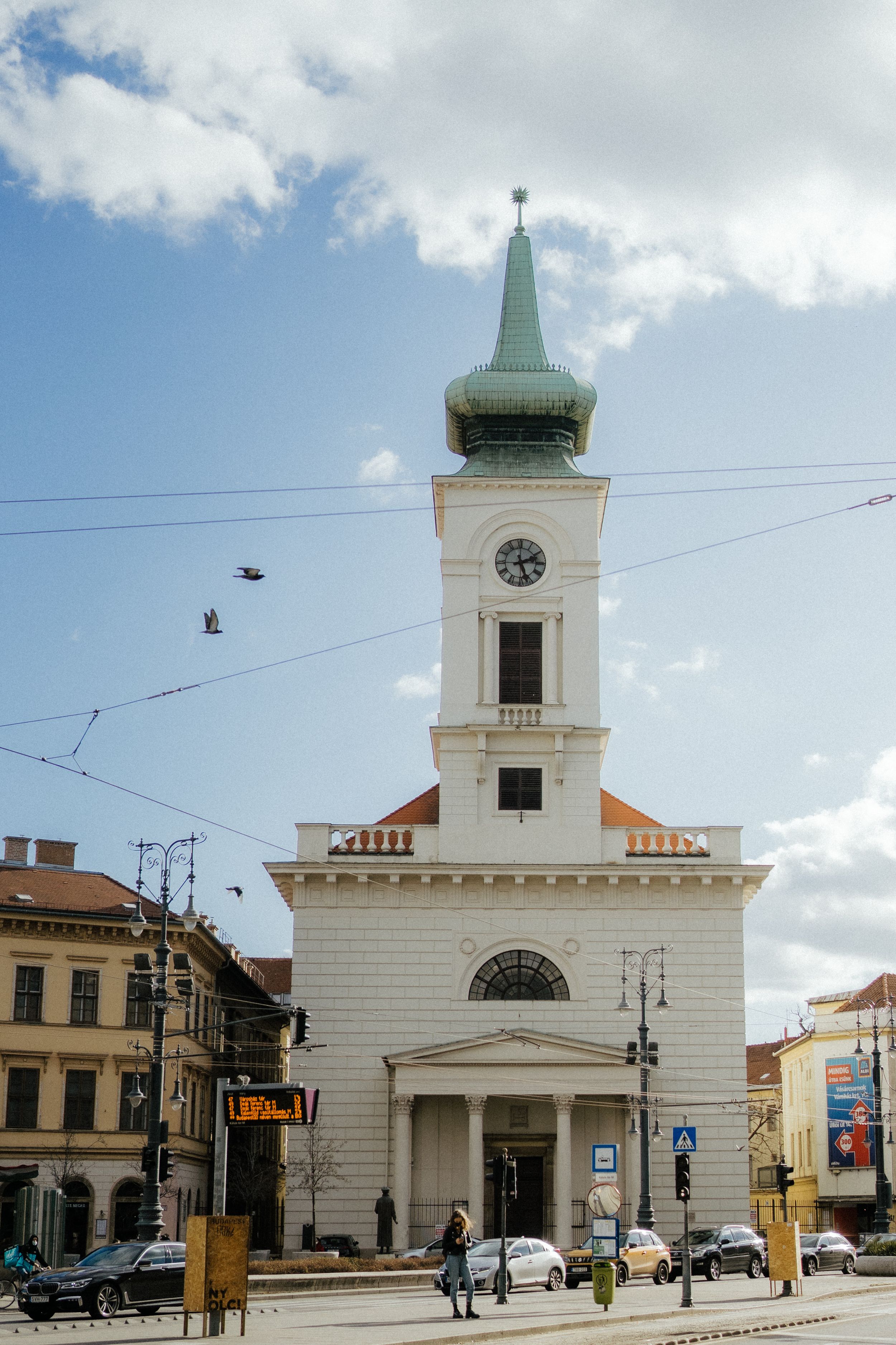 Historic white church with a tall green spire and clock tower stands prominently on a city street corner in Europe.