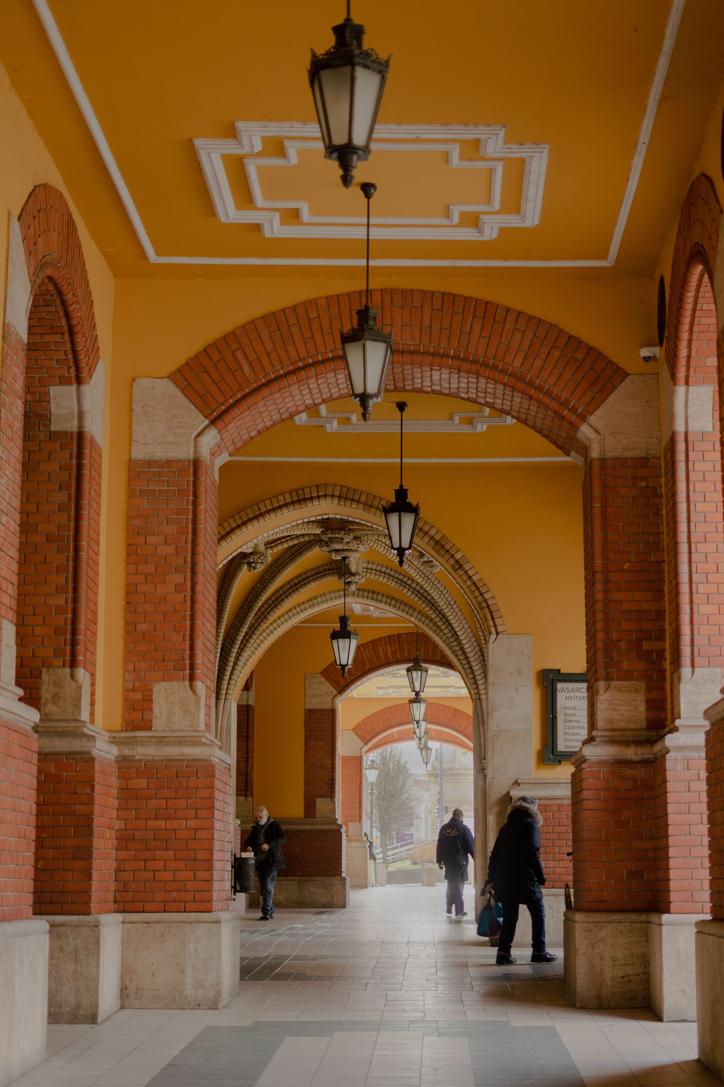 A yellow and brick archway corridor with hanging lanterns and geometric ceiling design leads to a distant opening.