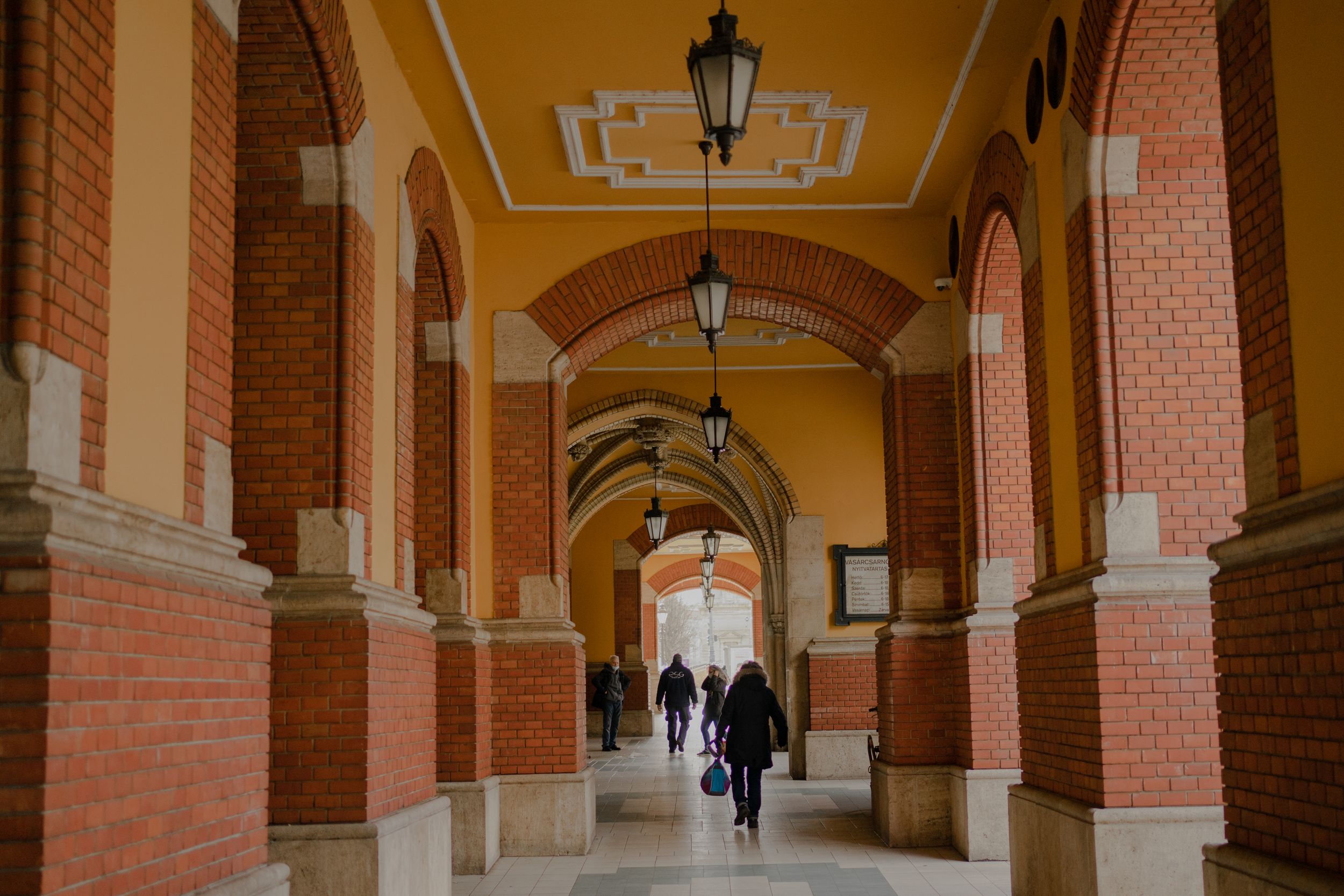 People walking down an ornate brick archway corridor with hanging lanterns and yellow-painted vaulted ceiling.