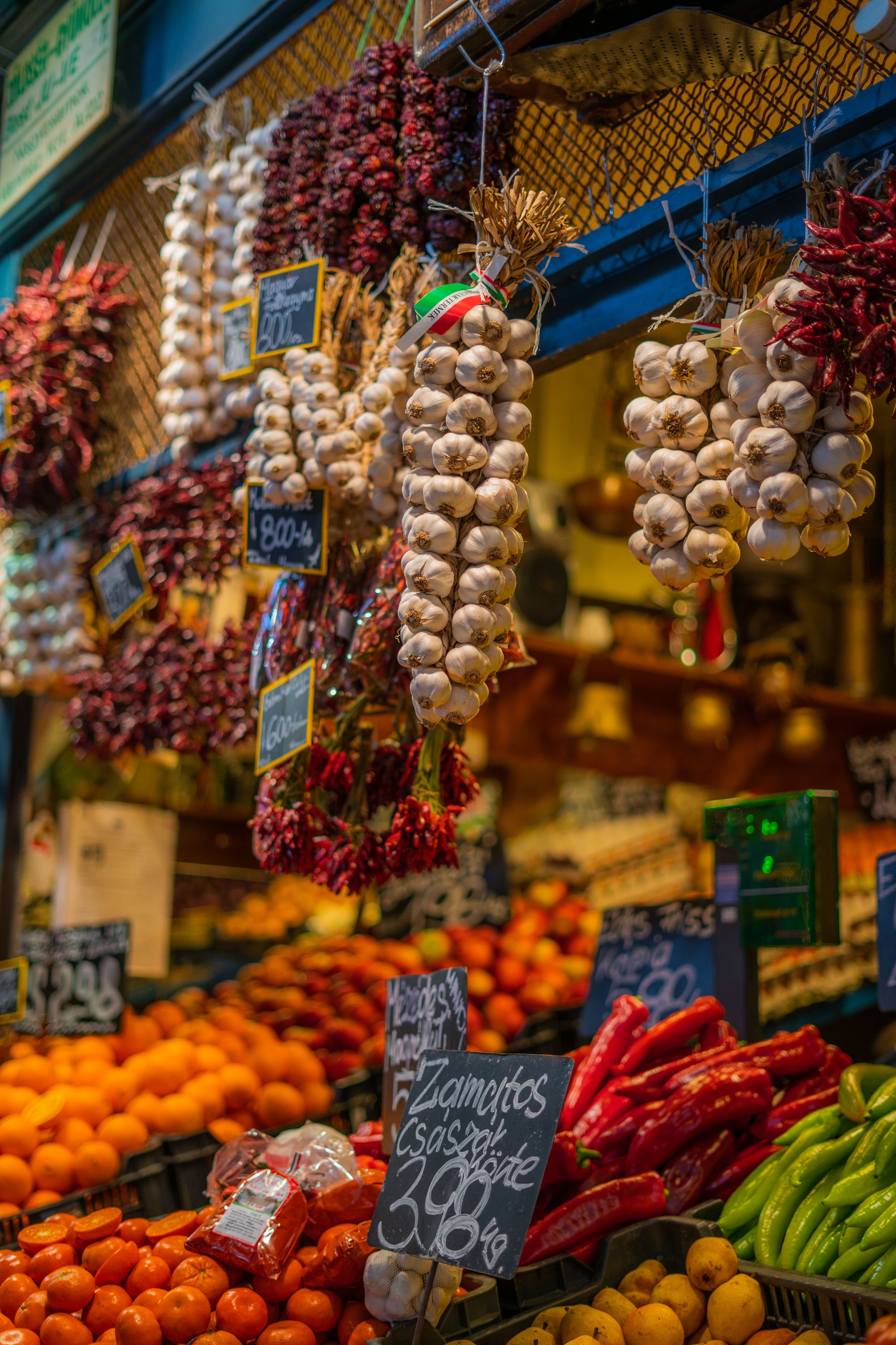 Fresh produce display at market with hanging garlic braids, colorful peppers, and oranges with vintage price signs.
