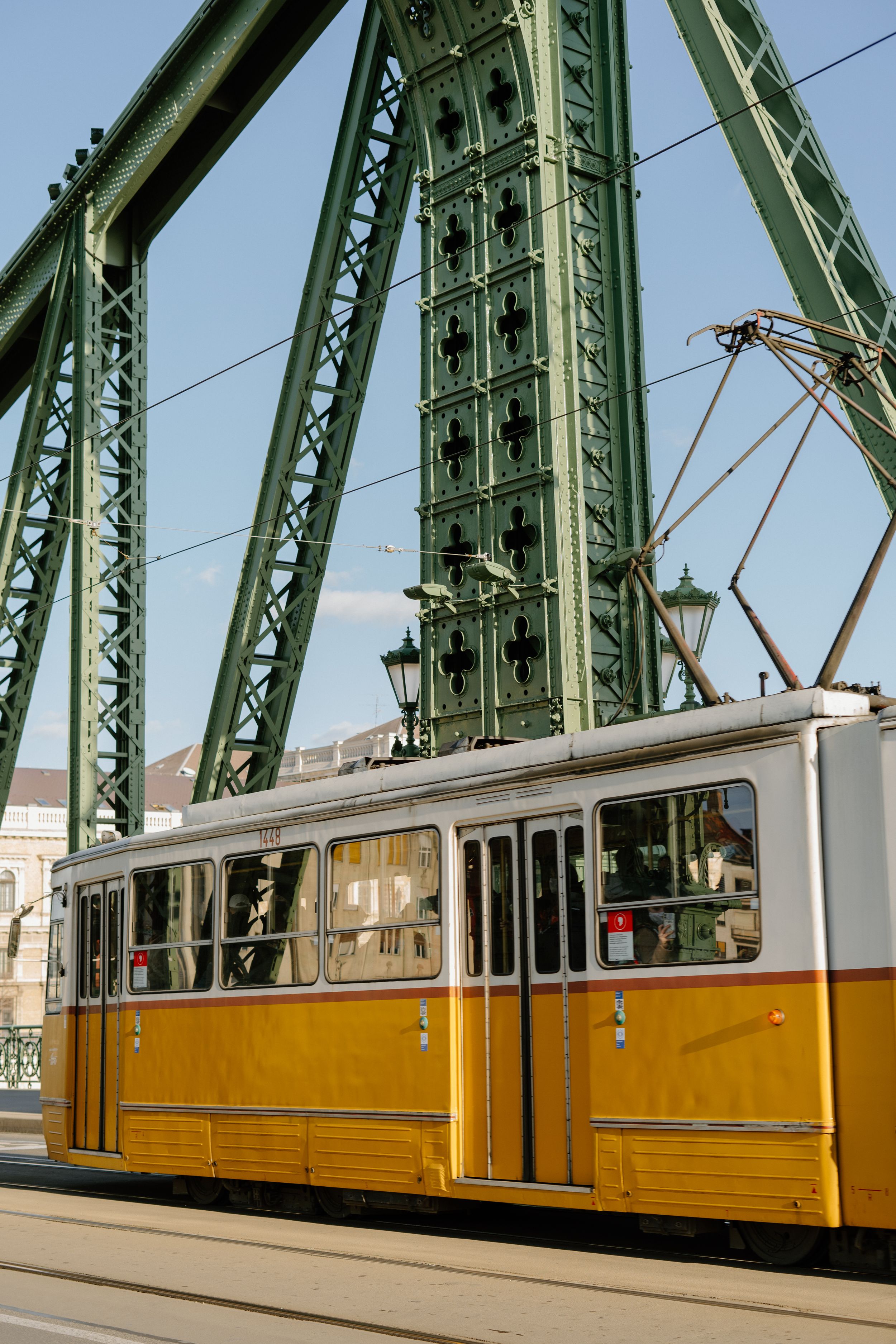 A vintage yellow and white tram passes under the green steel beams of Budapest's Liberty Bridge on a sunny day.