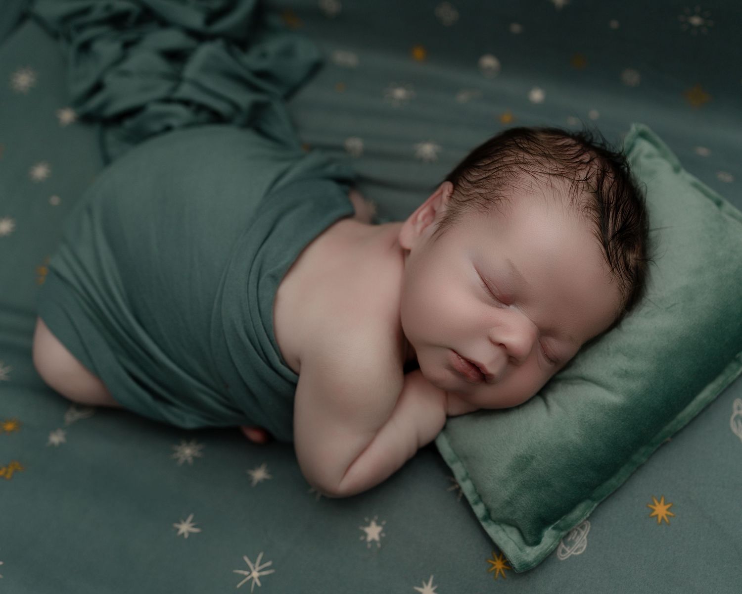 Sleeping newborn baby wrapped in teal cloth rests on teal pillow with gold star pattern background.