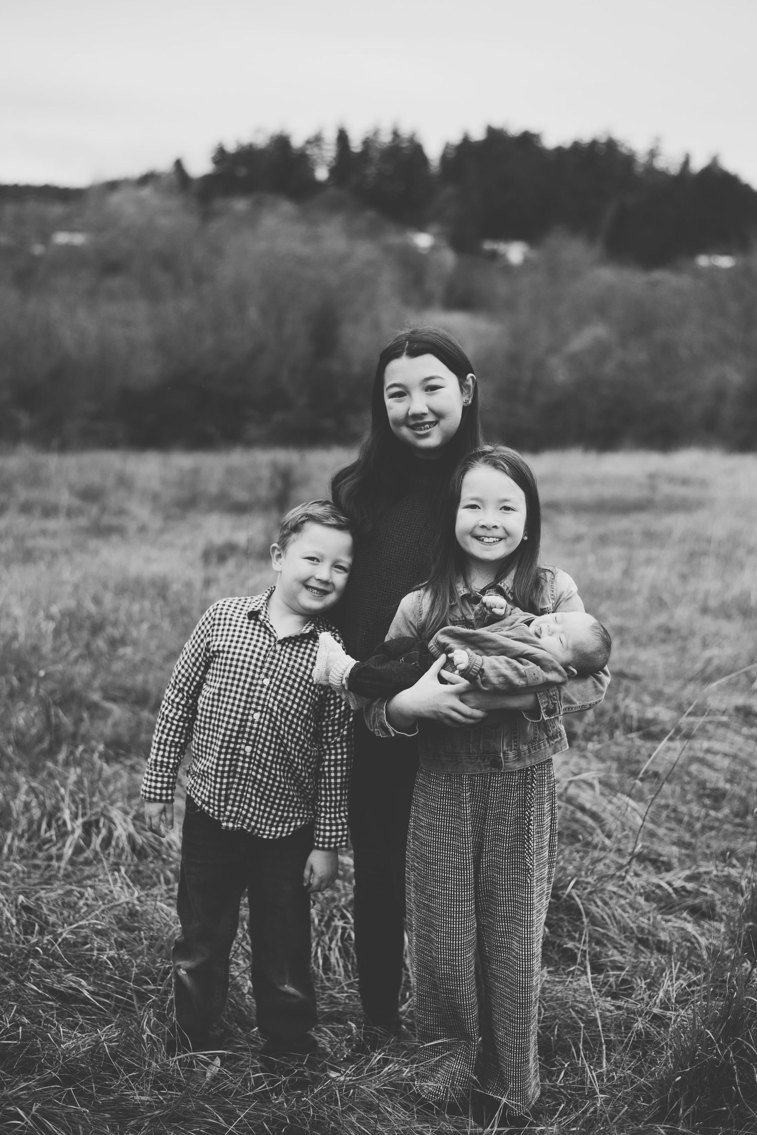 A black and white portrait of four siblings standing together in a grassy field with trees in the background.