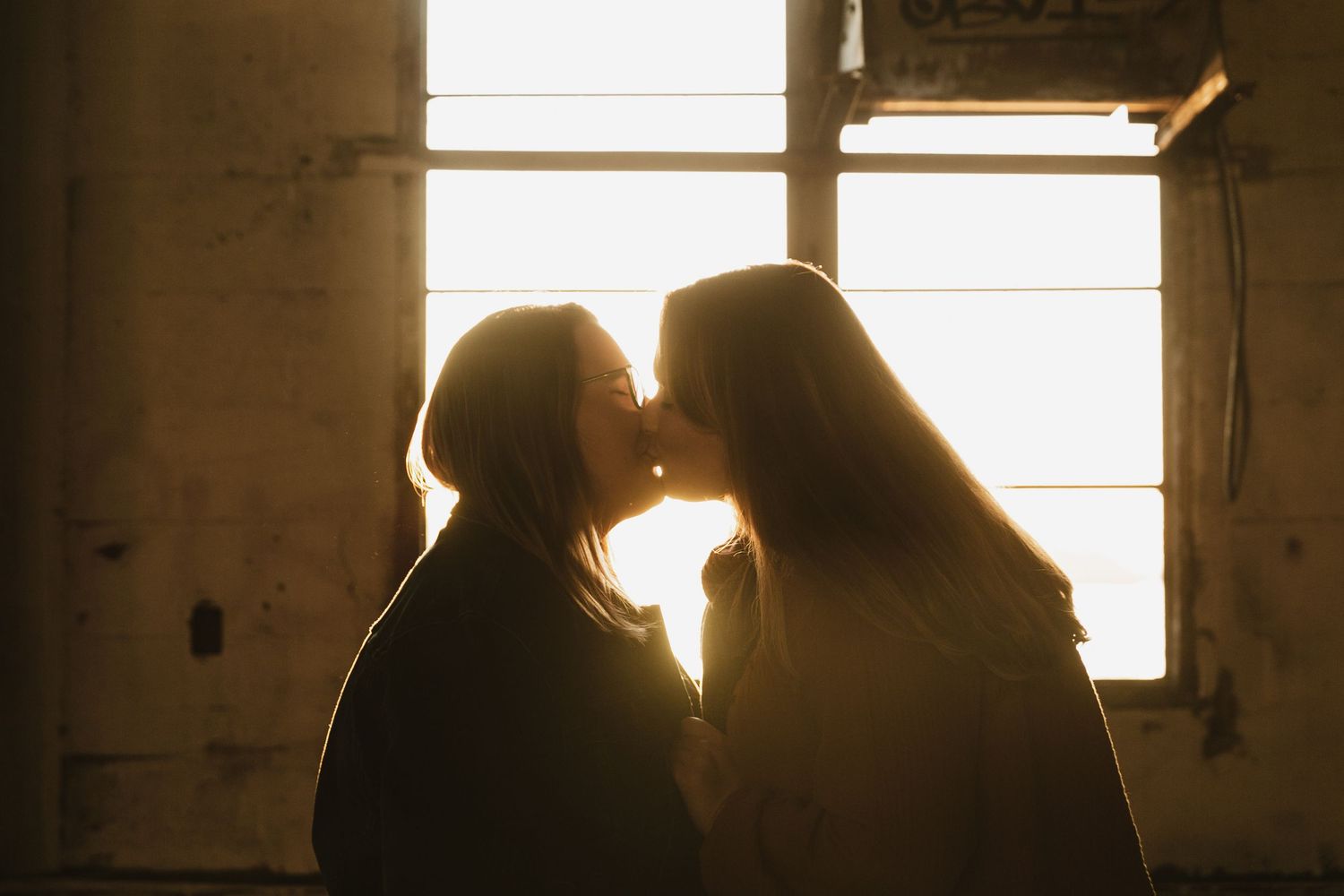 Two silhouettes share an intimate moment in front of a sunlit window creating a romantic backlit scene.
