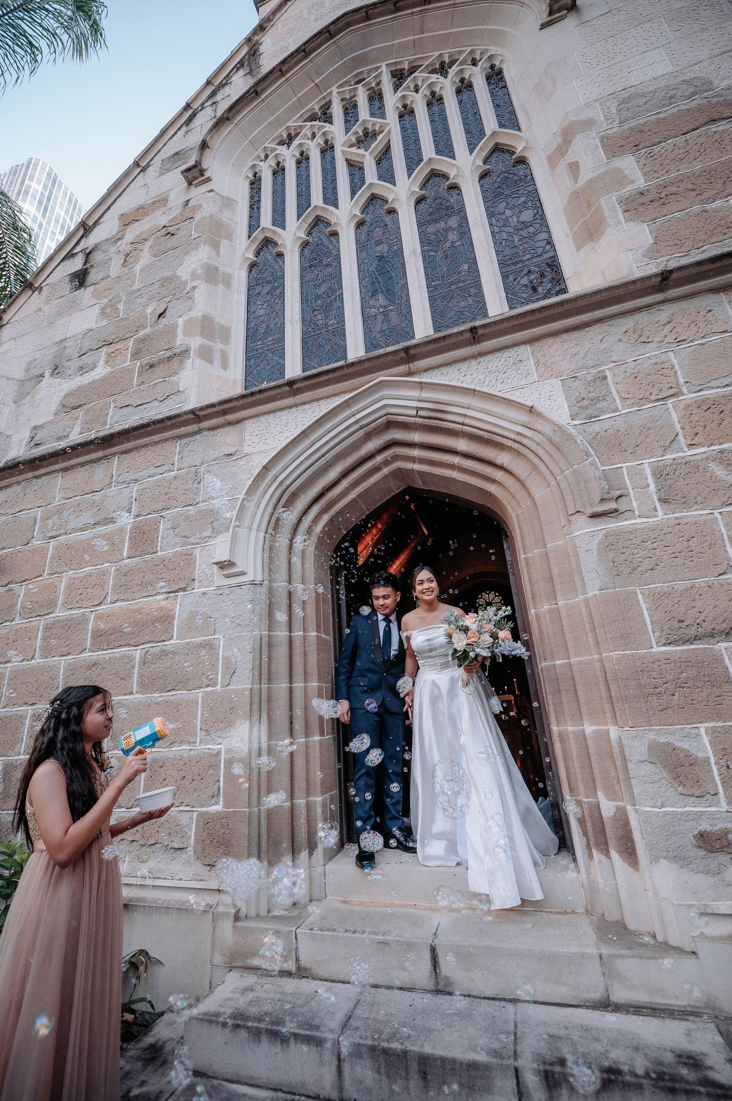 Newlyweds exiting gothic church doorway with guest blowing bubbles.