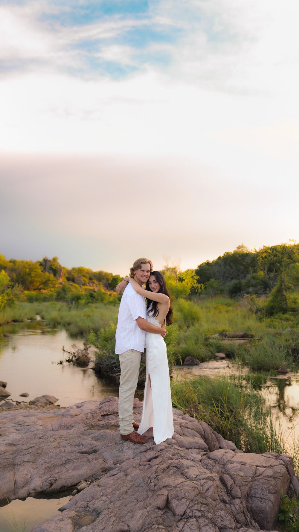 A couple in white outfits embrace on rocks by a serene river at sunset with lush greenery behind them.