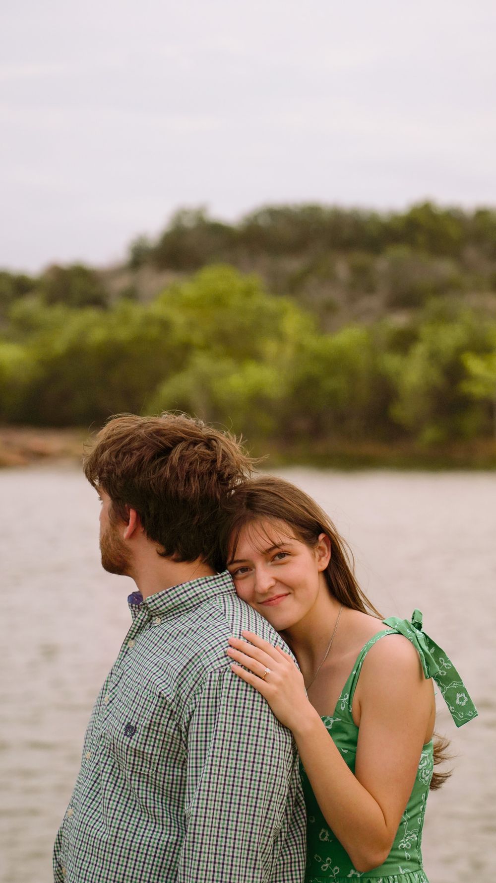 A smiling young woman in a green dress embraces a man near a lake with lush green trees in the background.