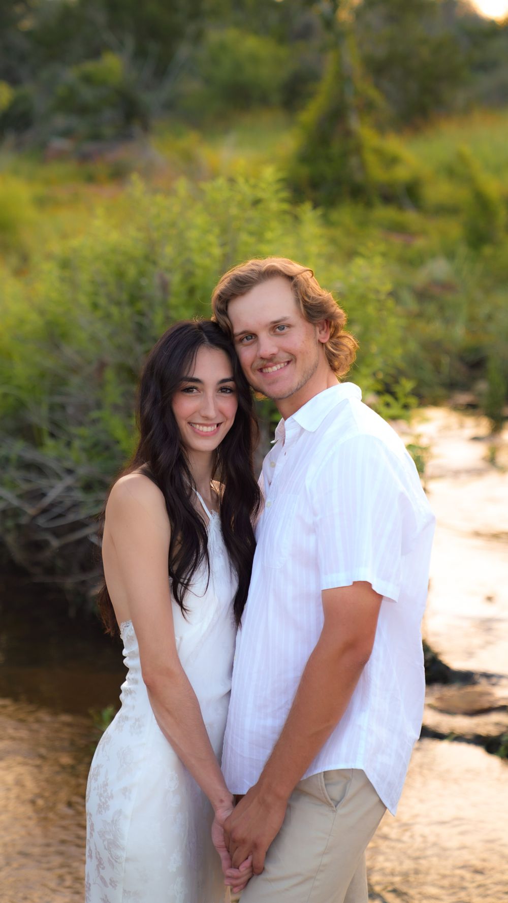 A smiling couple in white outfits poses together outdoors with lush green foliage in the background.