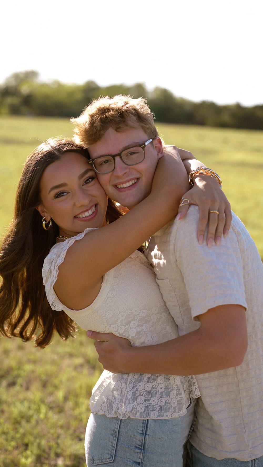 A smiling young couple embraces in a sunny green field, wearing casual white tops and jeans in a warm outdoor portrait.