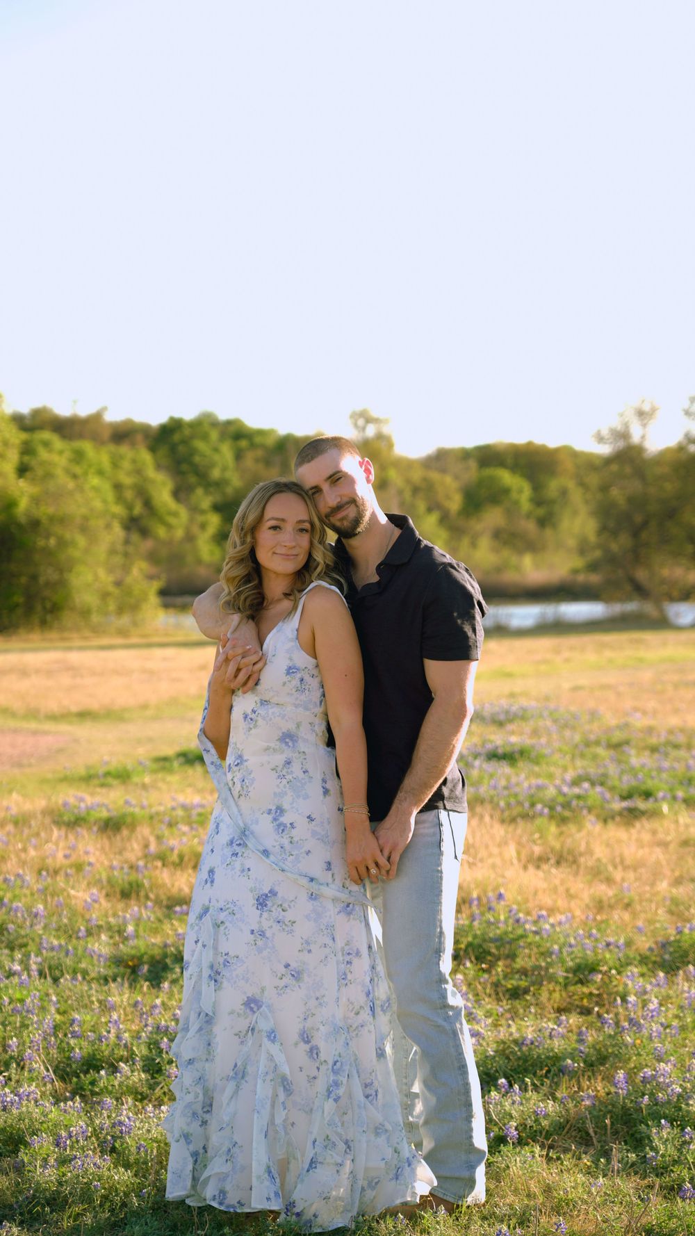 Couple posing in a sunny wildflower field, woman in floral dress, man in black polo, golden hour lighting.
