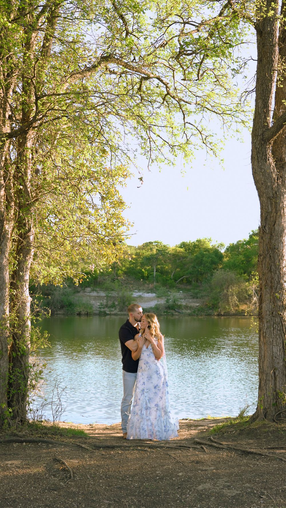 Couple embracing near a serene lake framed by tall trees in a romantic outdoor setting.