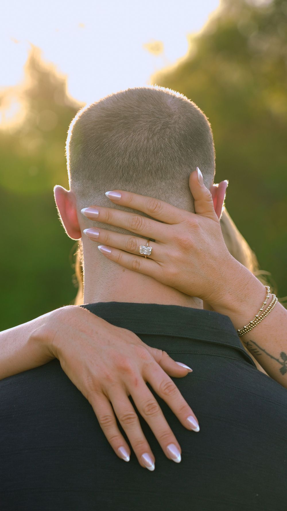 A woman with French manicured nails and an engagement ring embraces a man from behind in golden sunlight.