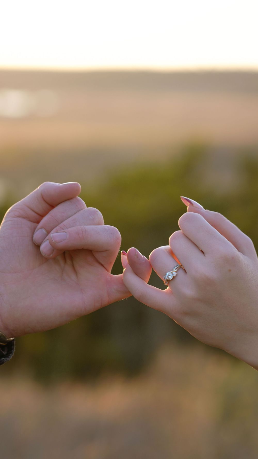 Two hands reaching towards each other with a diamond engagement ring glinting in the golden sunset light.