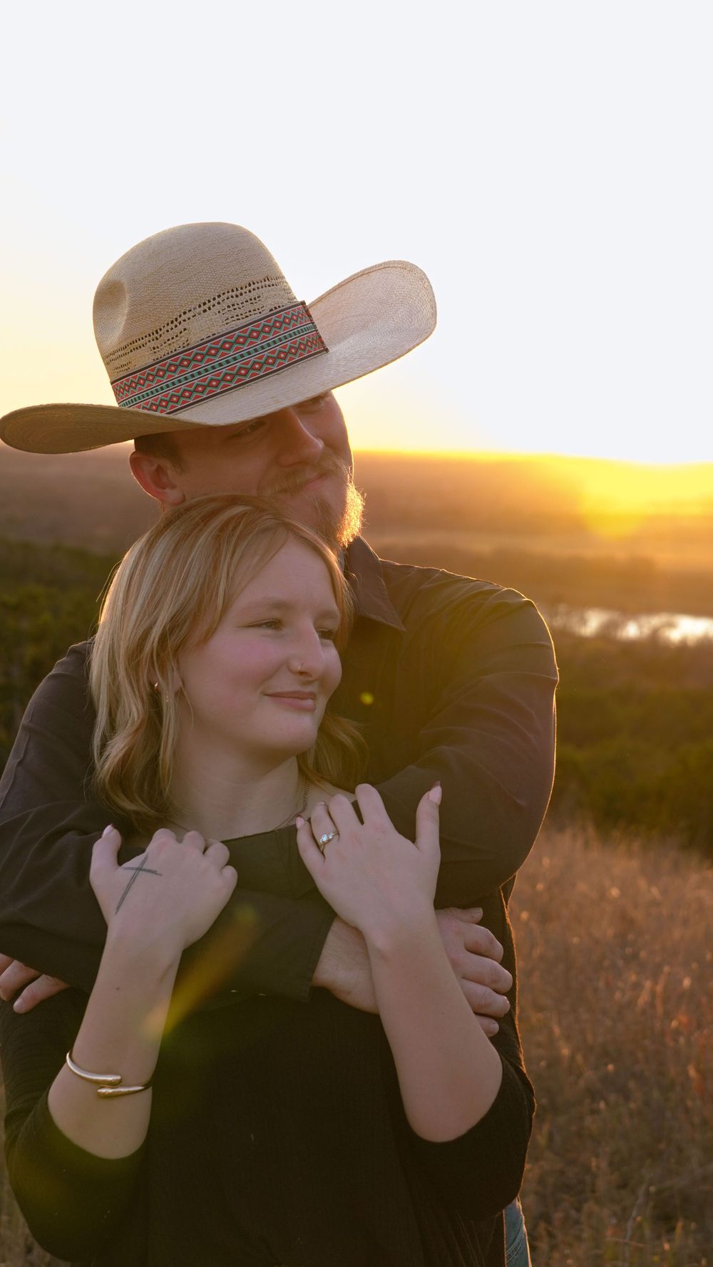A couple embraces in a sunlit field at golden hour wearing black clothing and a cowboy hat.