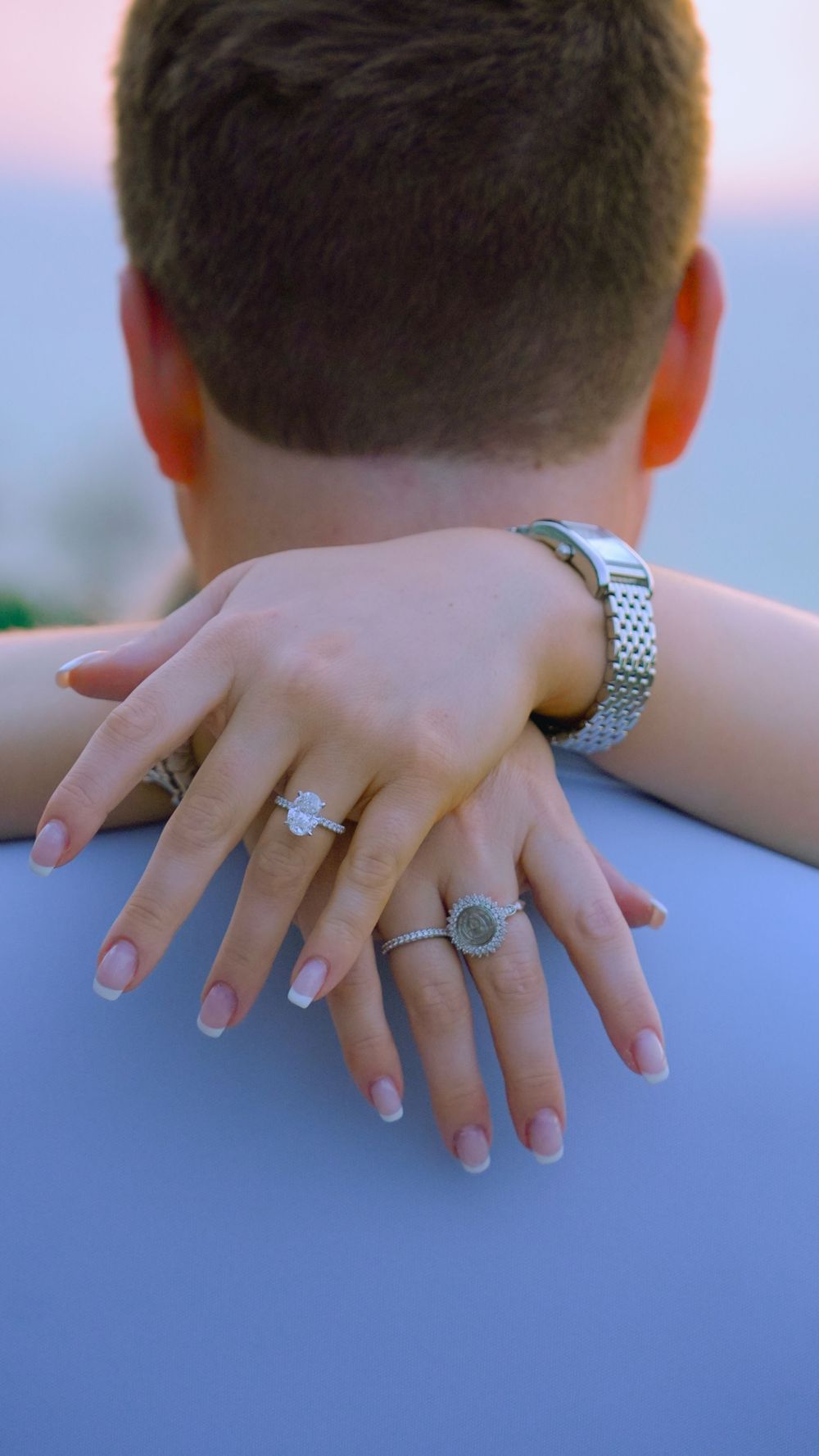 Hands with engagement rings and a silver watch resting on a surface during sunset with a soft pink sky in background.