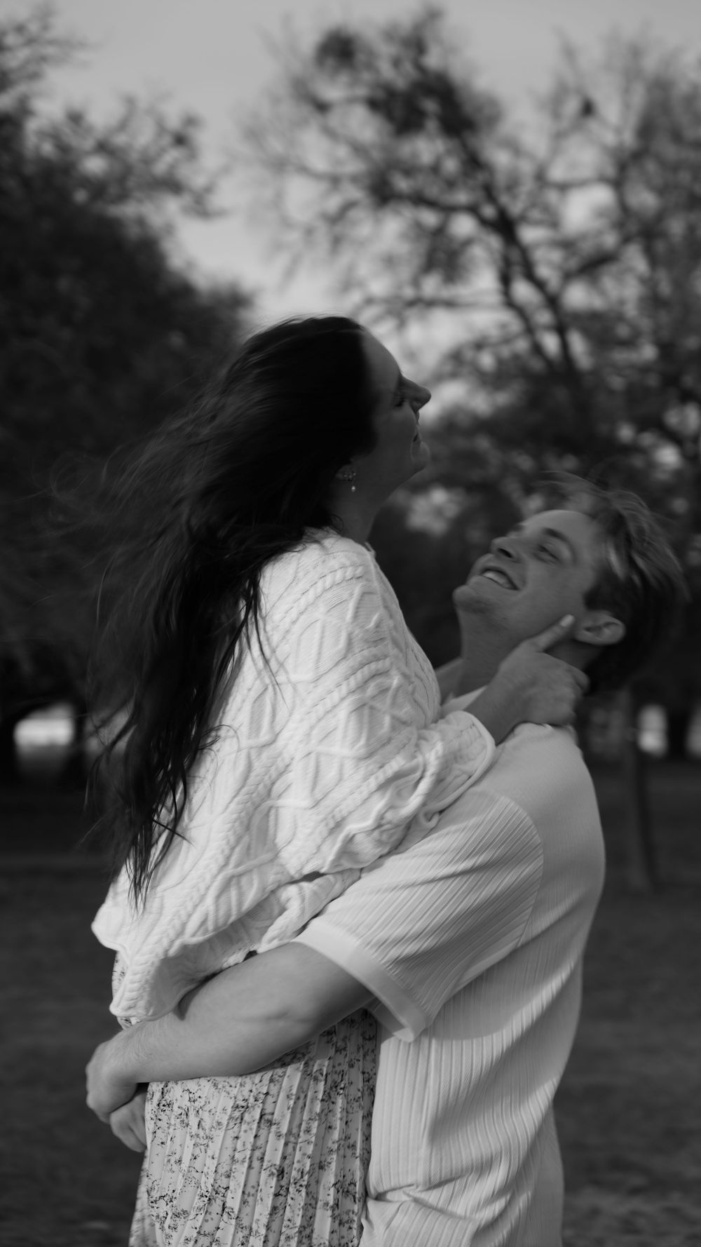A couple embraces joyfully outdoors in black and white, with the woman's long dark hair flowing in a candid moment.