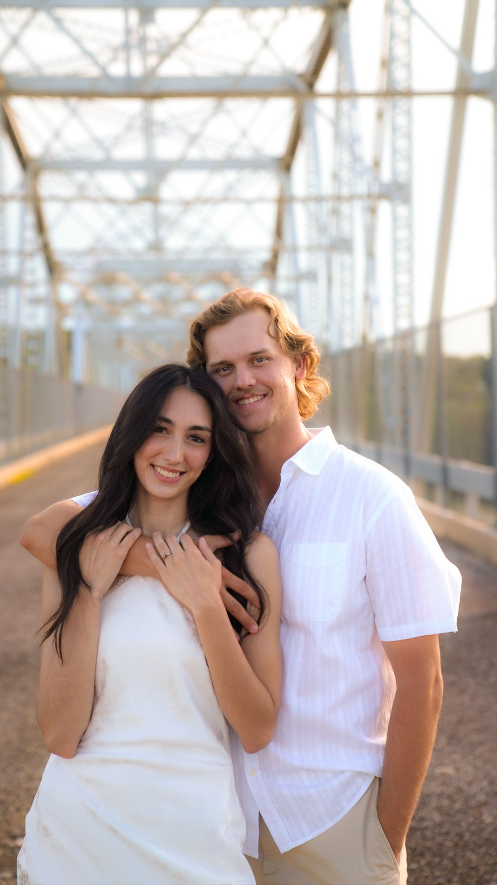 Smiling engaged couple posing together on bridge at golden hour in white outfits.