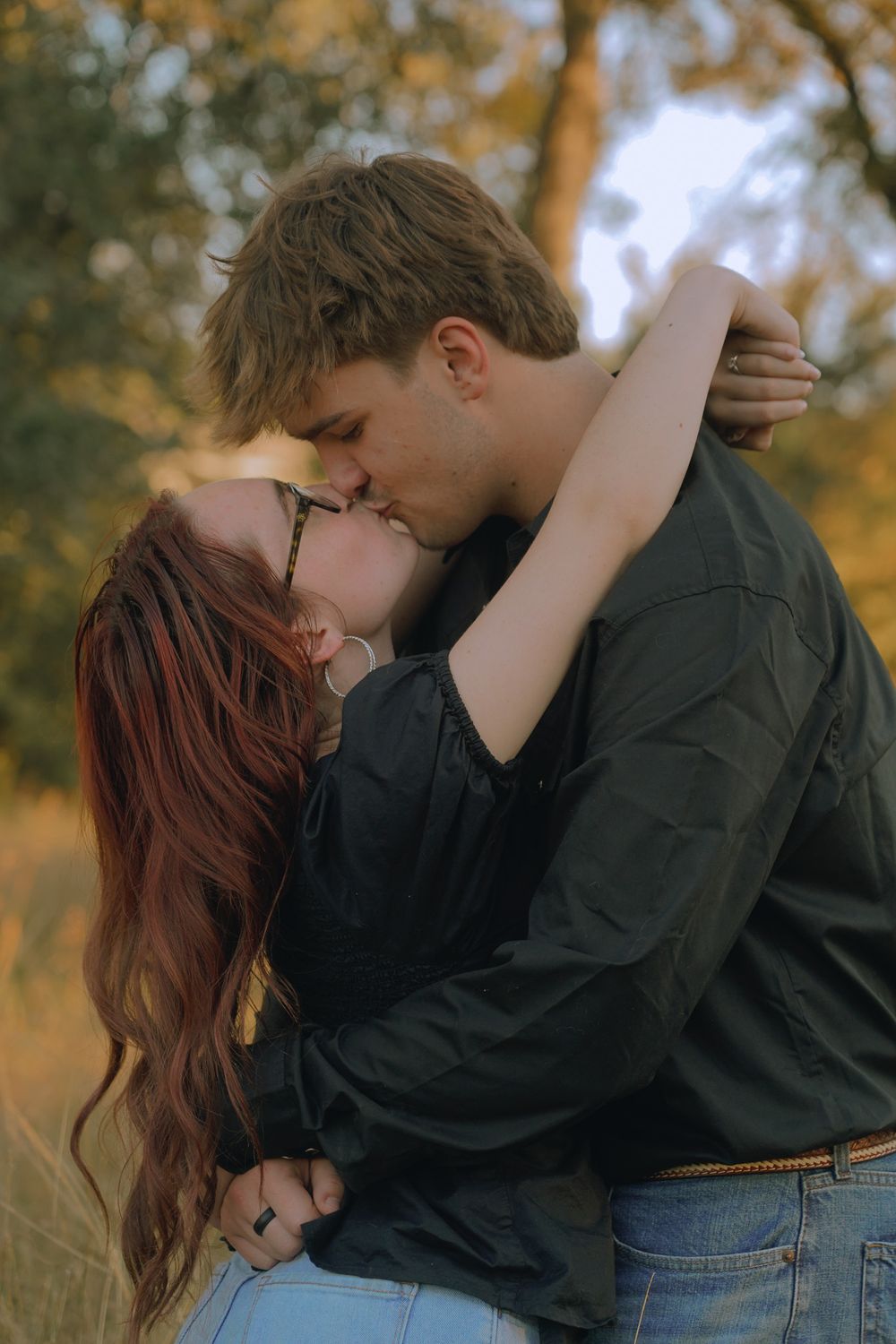 A romantic couple share a tender kiss outdoors during golden hour with autumn trees in the background.