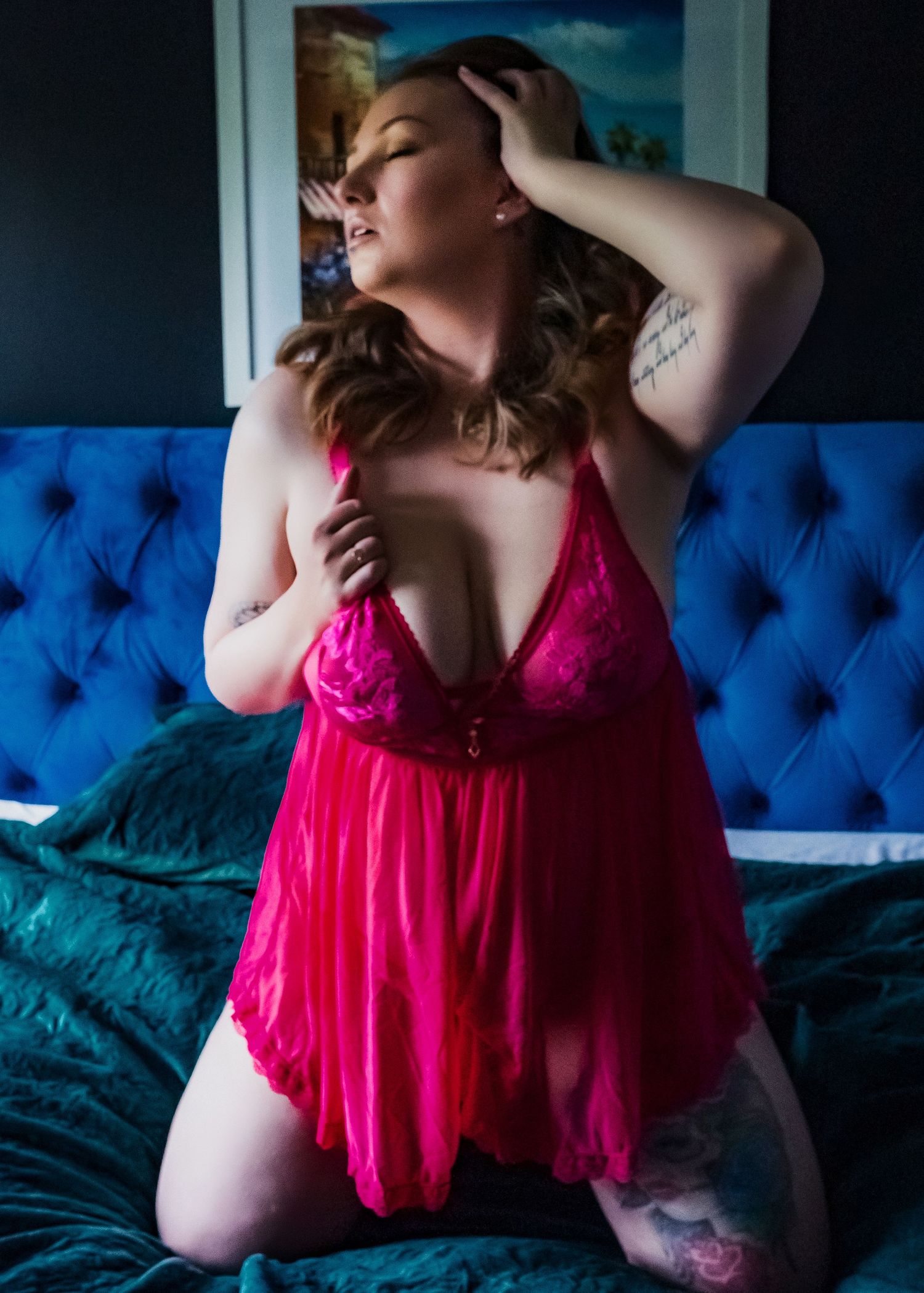Person in pink lace babydoll lingerie poses on blue tufted couch with decorative wall art in background.