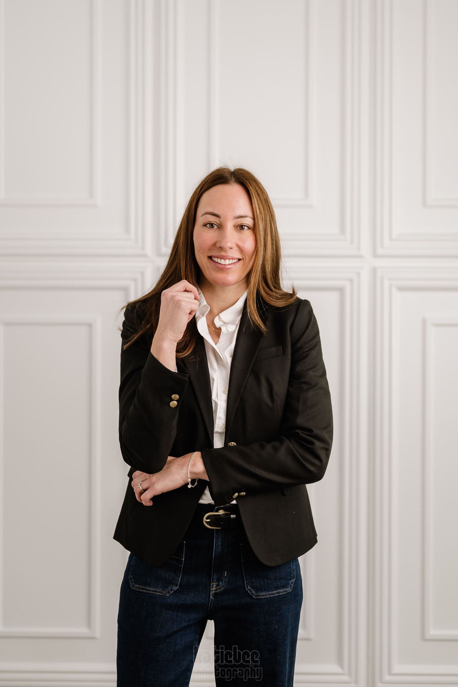 Professional portrait against white wall showing a business casually dressed person in black blazer and jeans smiling warmly.