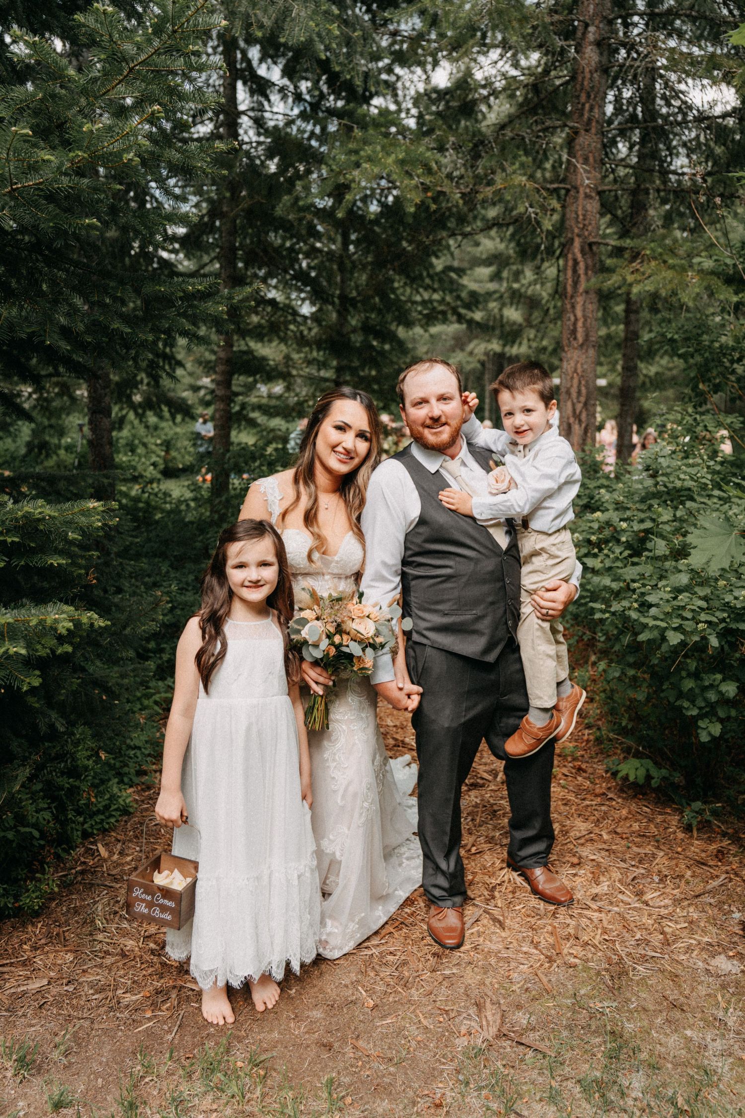 A family stands together on a forest path covered in pine needles surrounded by tall evergreen trees captured by wedding videographer Jovan
