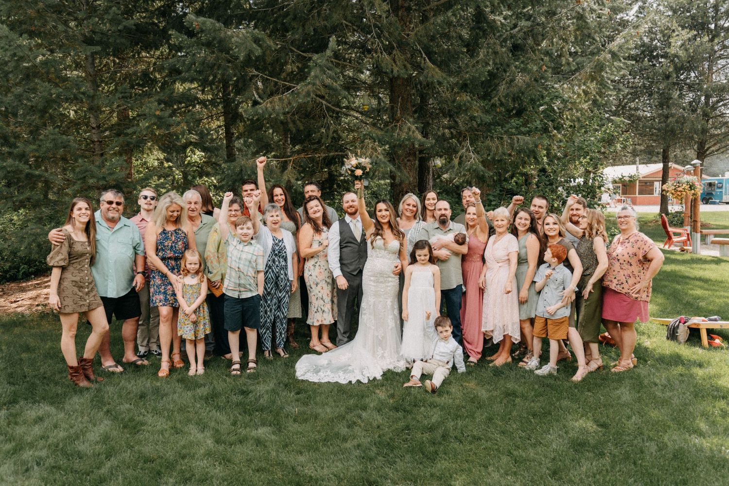 Large wedding party group poses on green lawn with evergreen trees in background captured by wedding videographer Jovan Films.