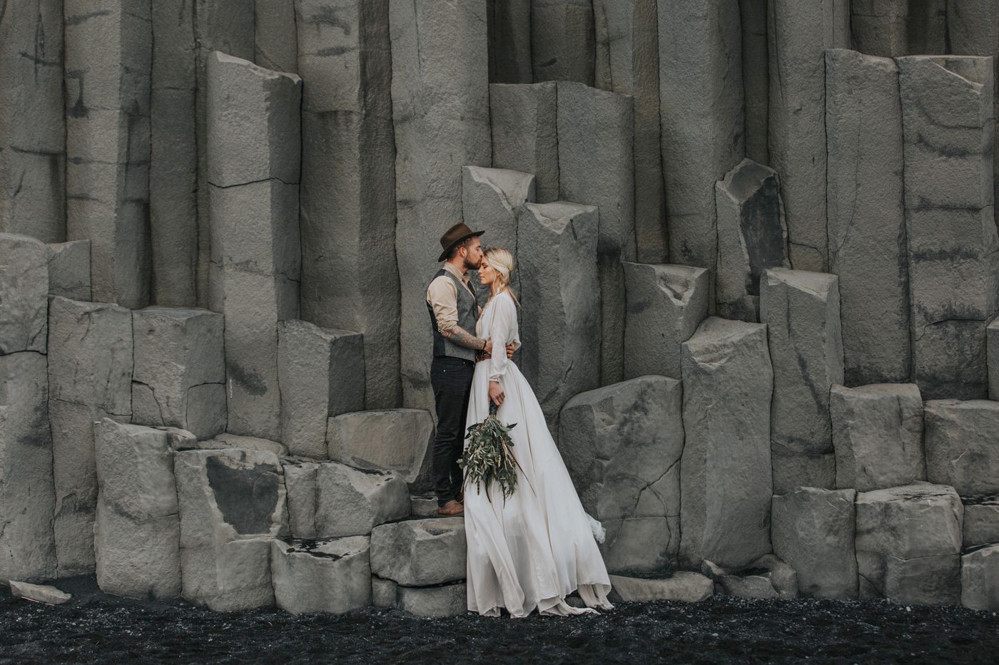 Romantic wedding couple embraces in front of dramatic basalt column cliffs at black sand beach in Iceland.
