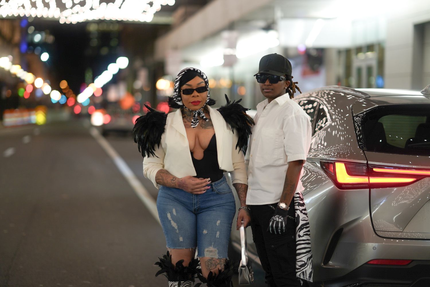 Fashion-forward couple posing against car in parking garage with statement accessories.