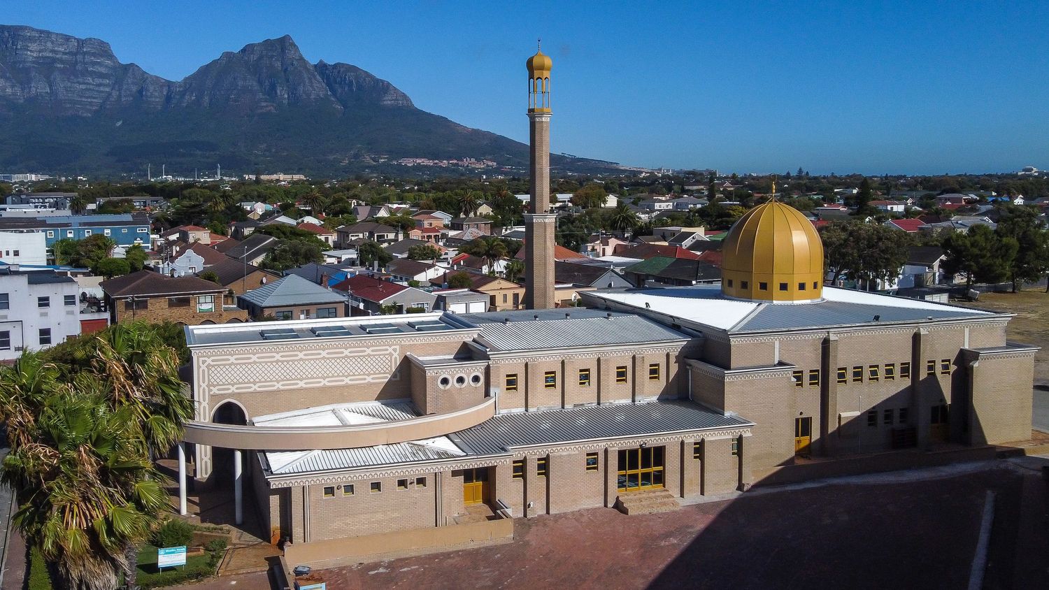 A modern mosque with golden dome and minaret against blue sky and mountain backdrop.