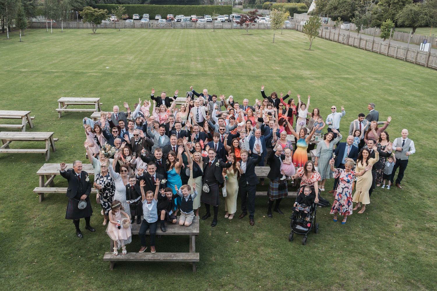 Large group of wedding guests gathered on wooden benches for an outdoor group photo on a green lawn.