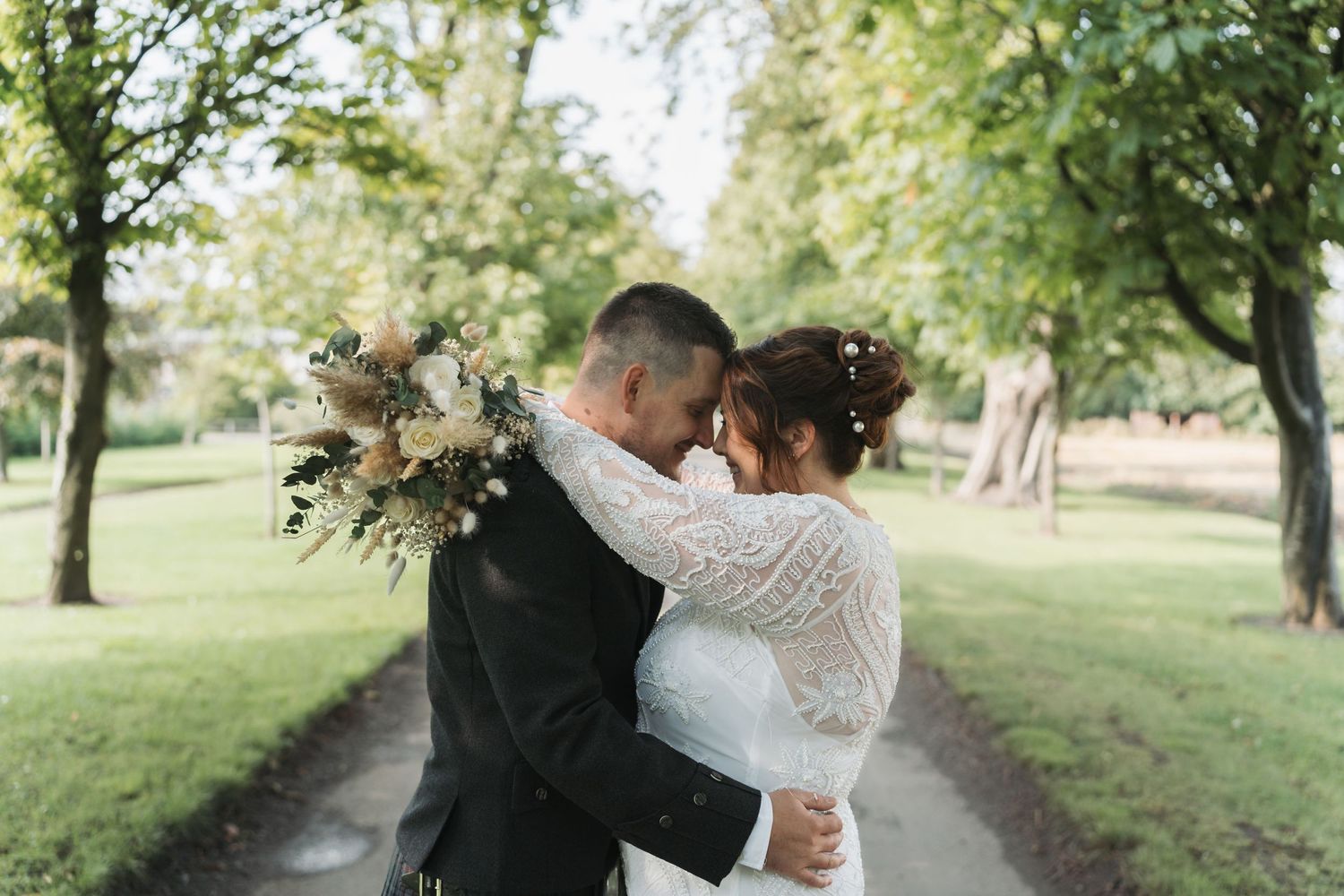 Newlywed couple embraces on a tree-lined path while the bride holds a rustic dried flower bouquet.
