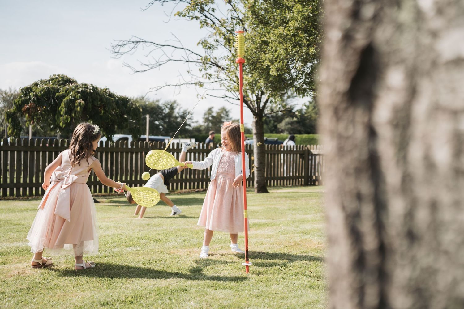 Children in pink dresses play badminton on a sunny lawn with a wooden fence in the background.