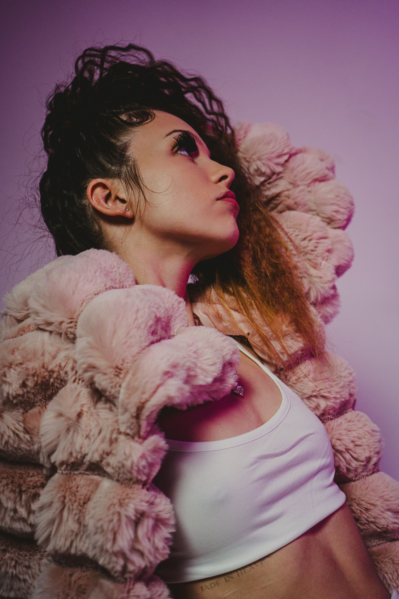 Artistic portrait against purple backdrop wearing pink fur coat and white crop top.