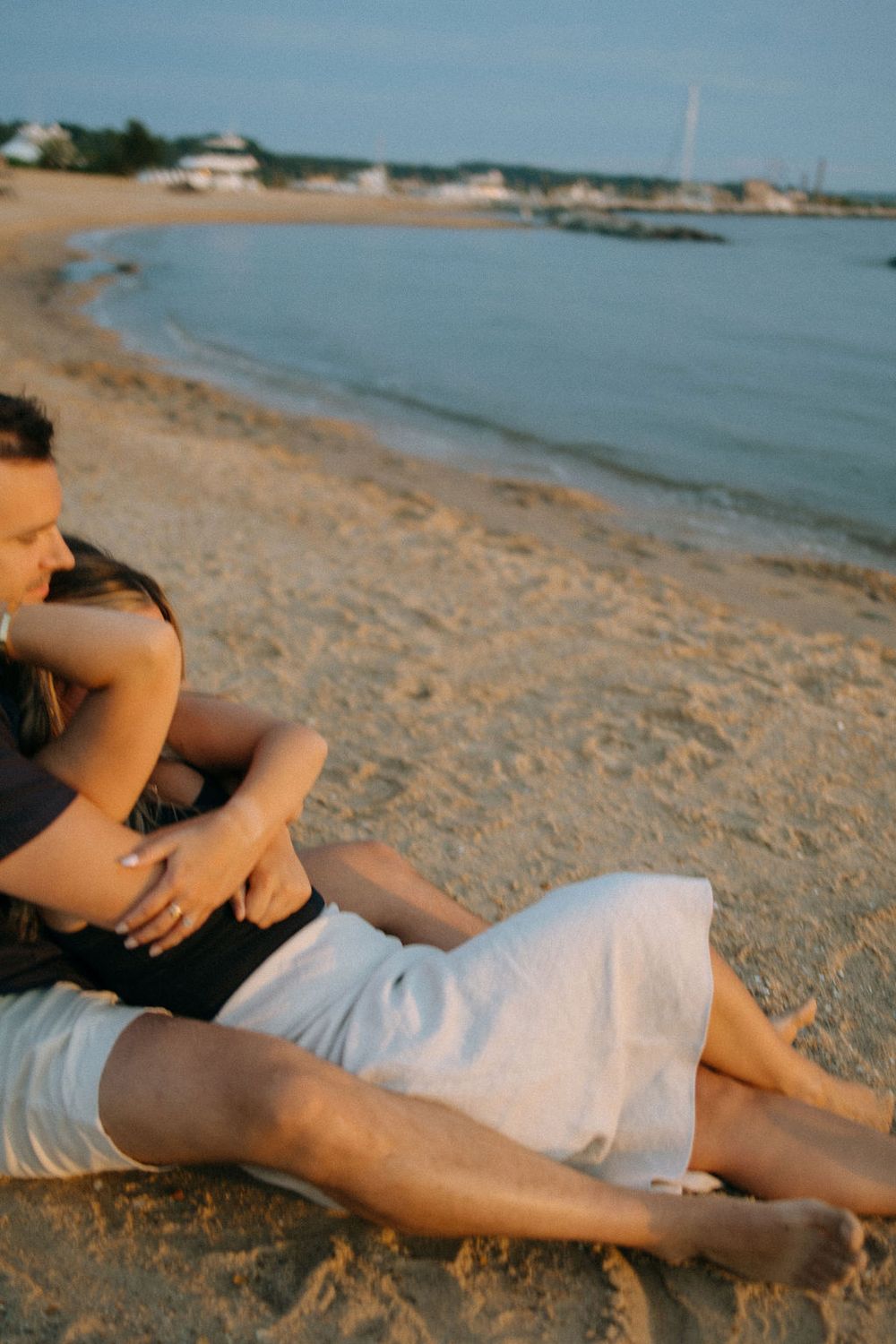 Couple sitting together on sandy beach at sunset, embracing near calm shoreline waters.