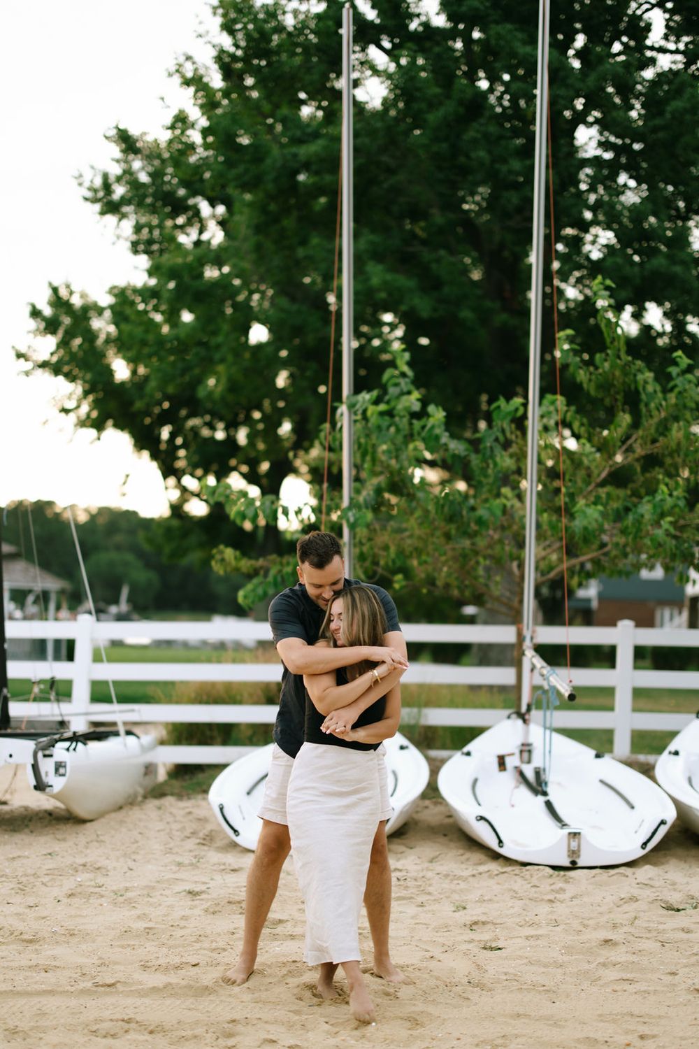 Couple embracing tenderly on sandy beach with white sailboats and green trees in background.