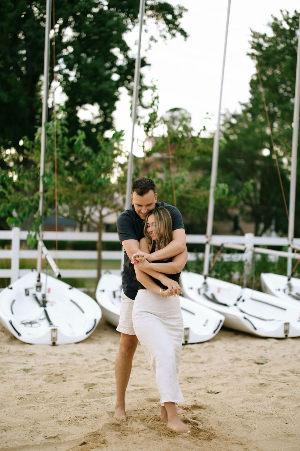 Couple embracing on sandy beach with white sailboats in background.
