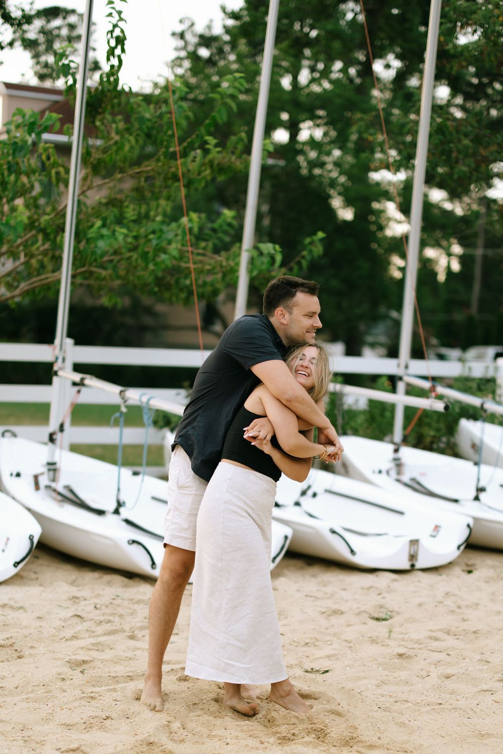 Man lifting woman in playful embrace on sandy beach with boats.