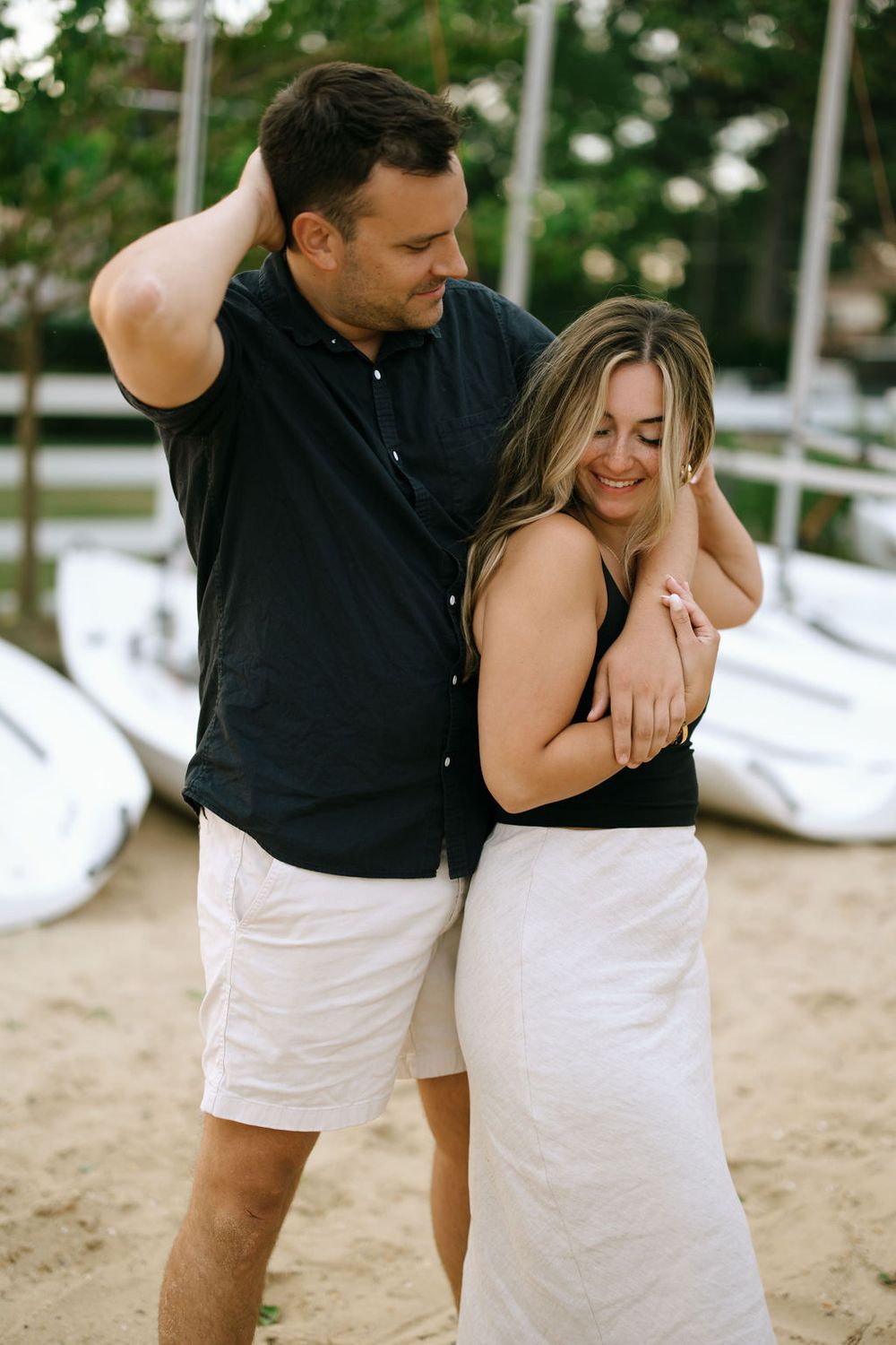 Happy couple laughing and embracing on beach near white sailboats.
