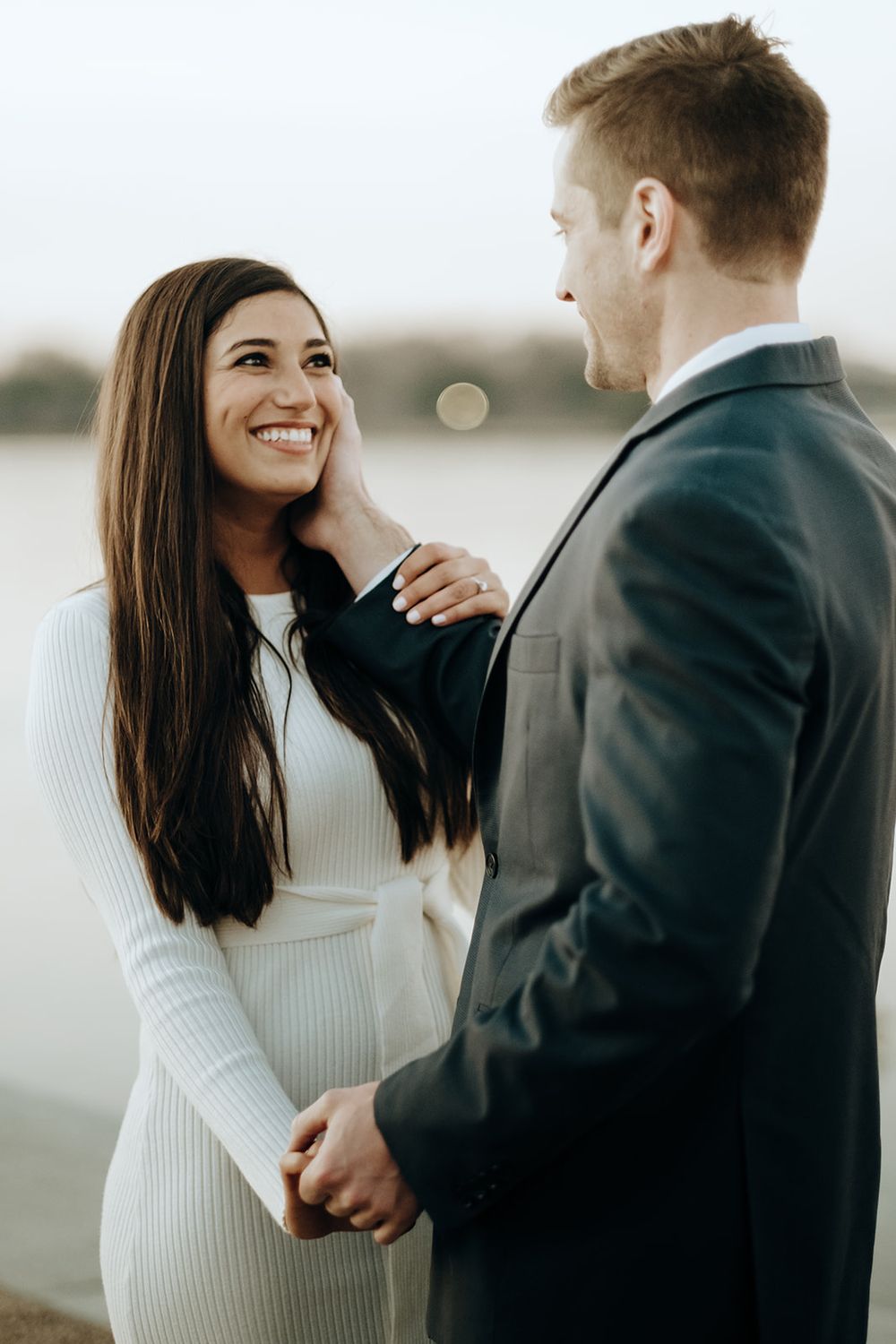 Man tenderly touches womans face as she smiles at him near a serene waterfront at dusk.