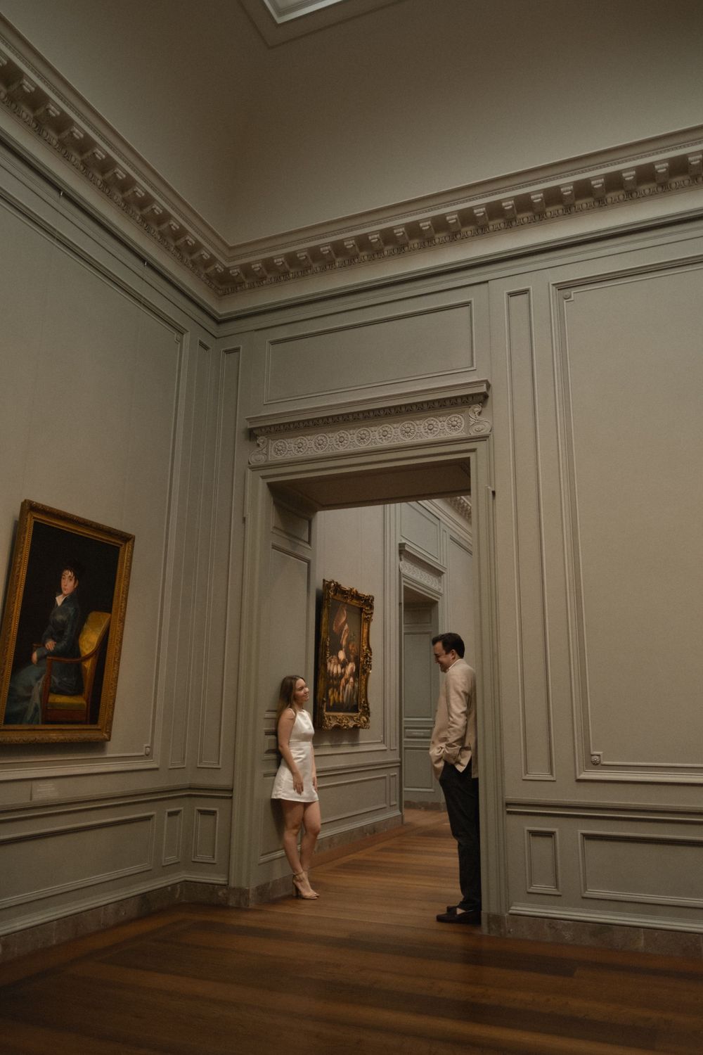 Couple conversing in an elegant art museum gallery with classic paintings and ornate moldings.