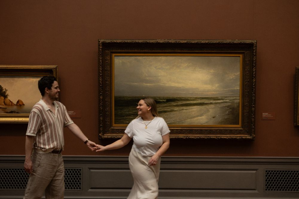 Smiling couple holding hands walking past a large seascape landscape painting on a terracotta museum wall.