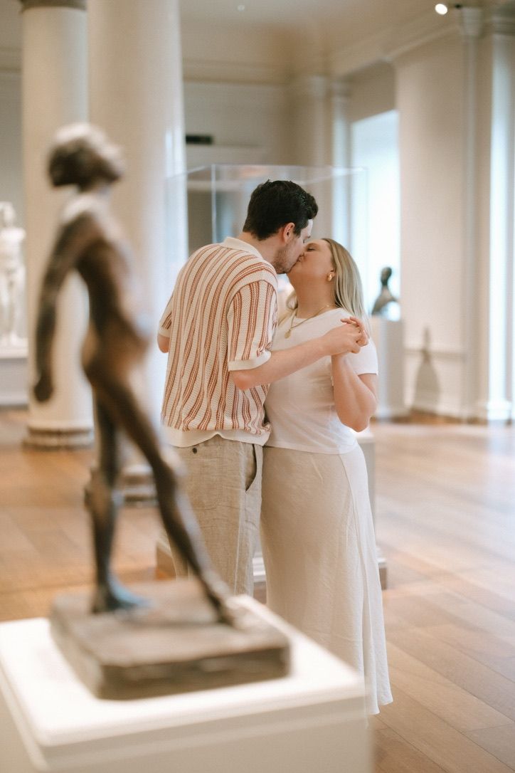 Couple sharing a tender kiss in a museum sculpture gallery with bronze statues nearby.