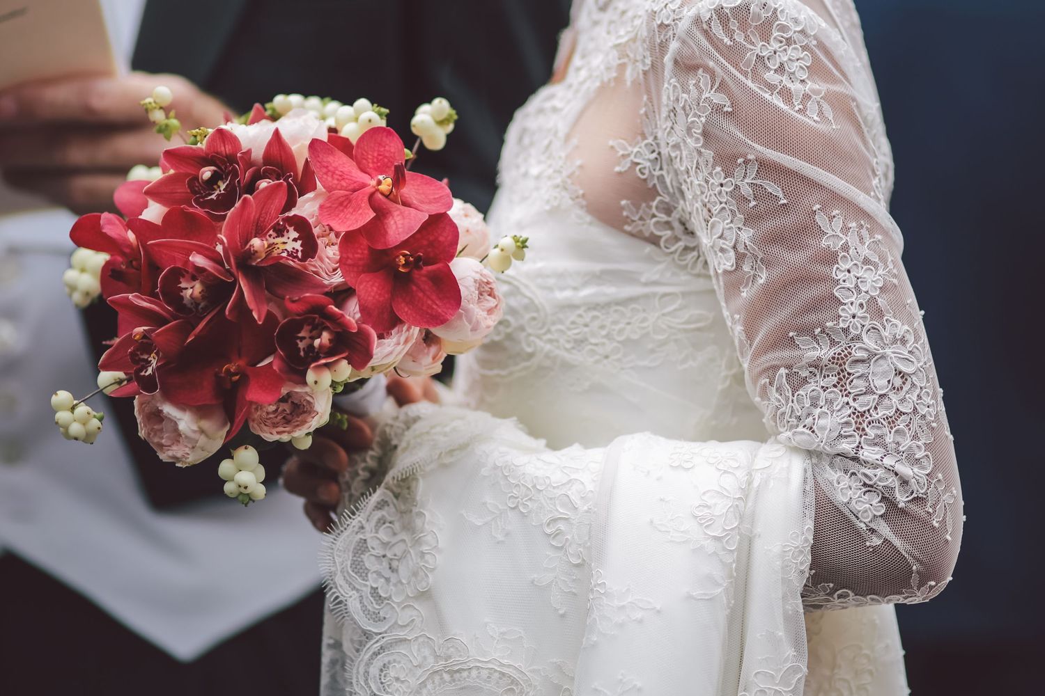 Deep red rose wedding bouquet held against white lace dress sleeves.