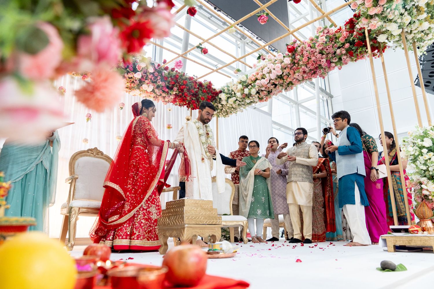 A couple in traditional attire performs a phera hindu under a floral mandap, surrounded by guests. The scene is vibrant and celebratory