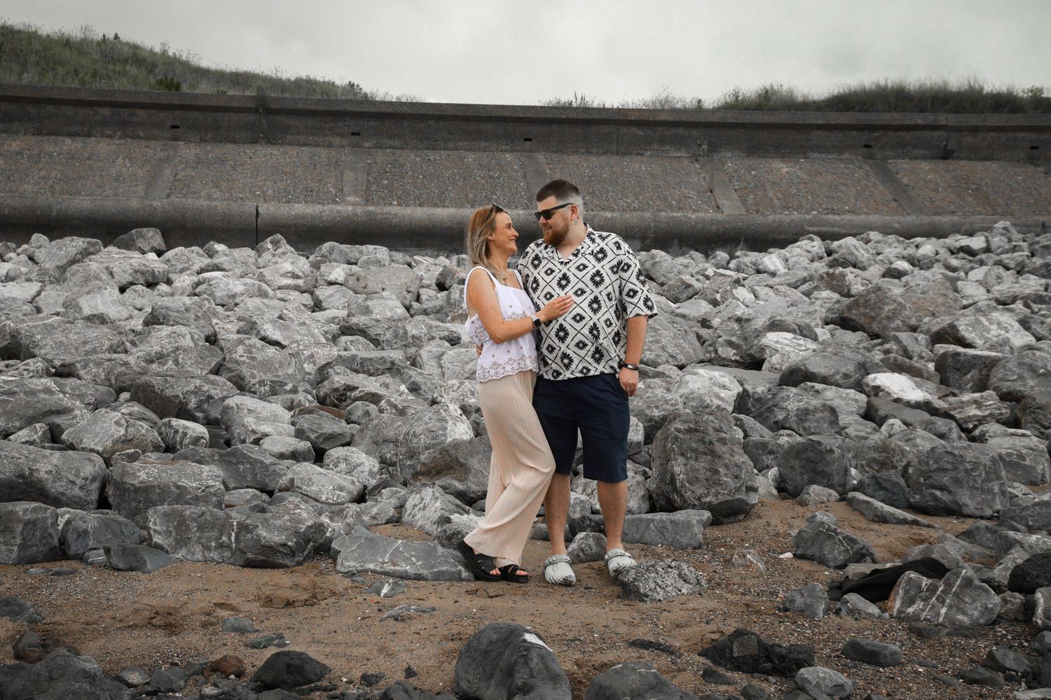 Couple embracing on rocky terrain near a lake during an overcast day.