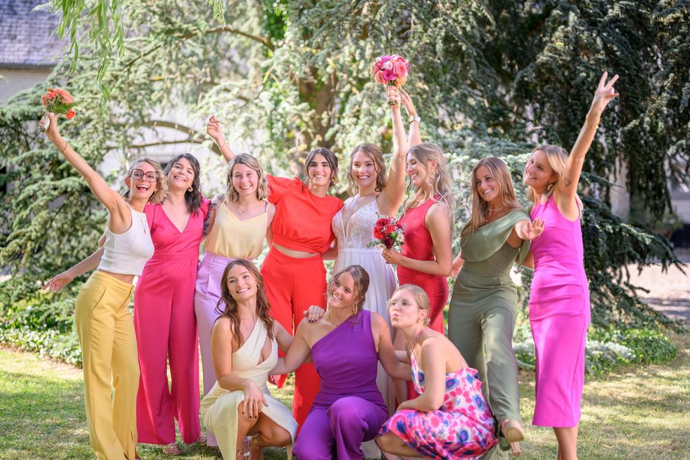 A group in colorful jumpsuits and dresses pose together joyfully with raised arms in a sunny garden setting.