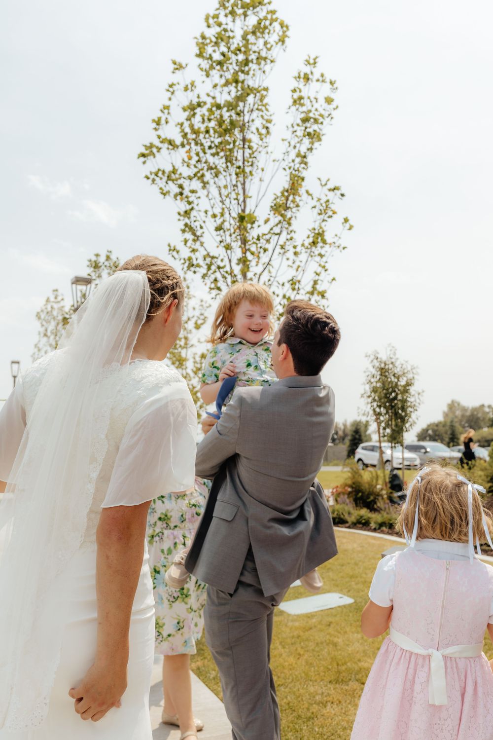 Groom in gray suit lifts a laughing child while bride in white dress stands nearby at outdoor wedding.